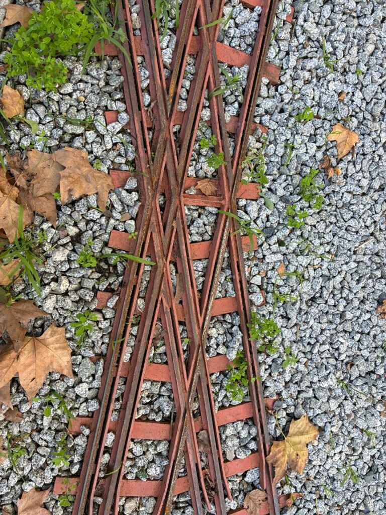 Estructura metálica oxidada sobre grava con hojas secas, formando un patrón geométrico en el suelo