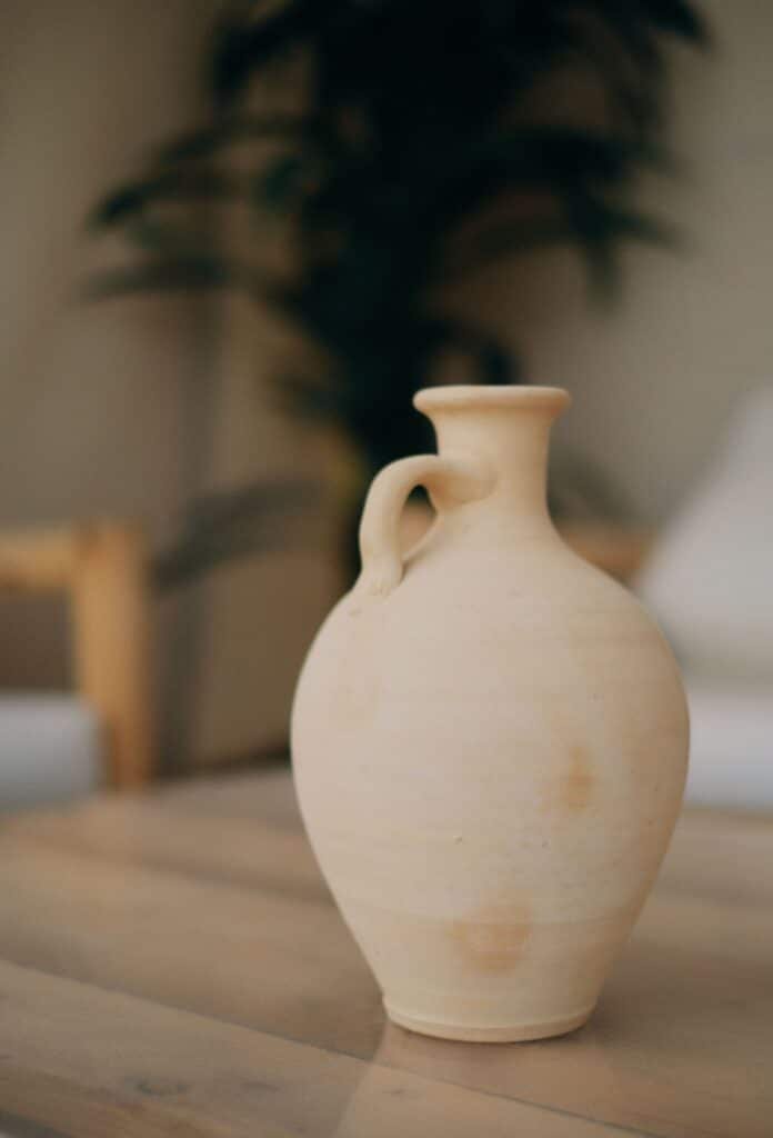 Jarrón de tono crema con un asa sobre una superficie de madera, con el fondo desenfocado.